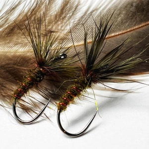 May include: Two fishing flies with curved hooks, featuring olive green bodies, dark brown feather wings, and red thread accents. A brown feather is in the background.