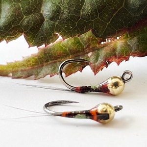 May include: Two fishing flies with curved hooks and gold-colored heads. The bodies of the flies are painted with a mix of red, green, and orange colors. A green and red leaf is in the background.