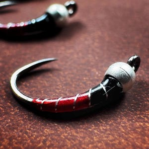 May include: Close-up of a fishing hook with a curved design. The hook features a silver eye and a body that transitions from black to red, with silver accents. The hook is designed for fishing.