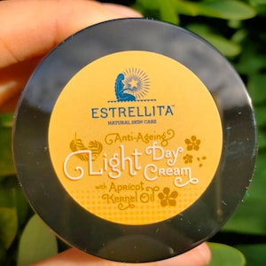 May include: A black jar of Estrellita Natural Skin Care Light Day Cream with apricot kernel oil. The cream's label is yellow with the brand name and product details in white and blue. The jar is held in a hand.