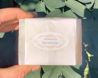 May include: A bar of handmade soap wrapped in white paper. The soap is labeled with the text "ESTRELLITA Hand Made Soaps" in an oval design with gold accents. The soap is held in a hand, with green leaves in the background.