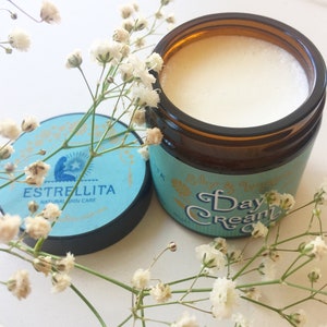 May include: A glass jar of white cream with a blue and gold label that reads "Estrellita Natural Skin Care" and "Day Cream". The jar is sitting on a white surface with white flowers.