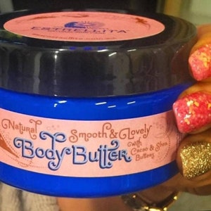 May include: A blue jar of body butter with a black lid and a pink label. The label reads "Natural Smooth & Lovely Body Butter" with the words "with Cacao & Shea Butters" below. The jar is held by a person with glittery nails.