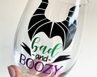 May include: A clear glass wine glass with a black silhouette of Maleficent's horns and the text "bad -and- boozy" in green and purple.
