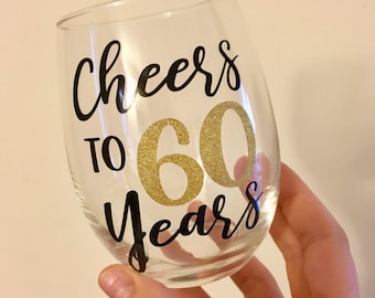 May include: Clear glass wine glass with black lettering that says "Cheers to 60 Years" with a gold glitter 60.