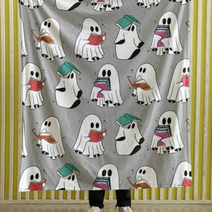 May include: A gray fleece blanket with a repeating pattern of white ghosts reading books. The ghosts are holding books of various colors and sizes. The blanket is being held up by a person's hands.