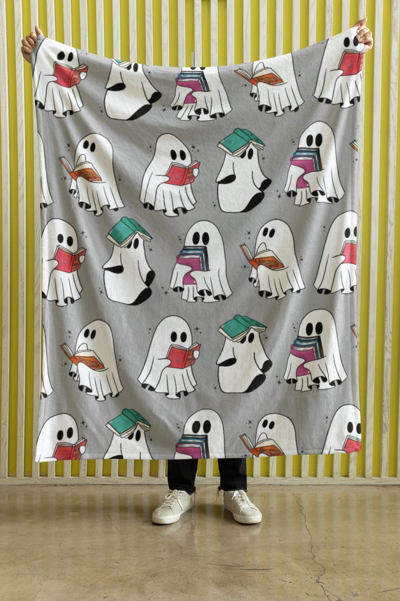 May include: A gray fleece blanket with a repeating pattern of white ghosts reading books. The ghosts are holding books of various colors and sizes. The blanket is being held up by a person's hands.