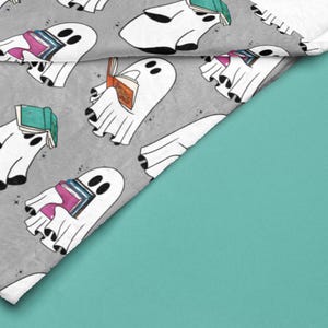 May include: A gray and white fabric with a repeating pattern of white ghosts holding stacks of colorful books. The ghosts are outlined in black and have small black eyes. The fabric has a subtle sparkle.