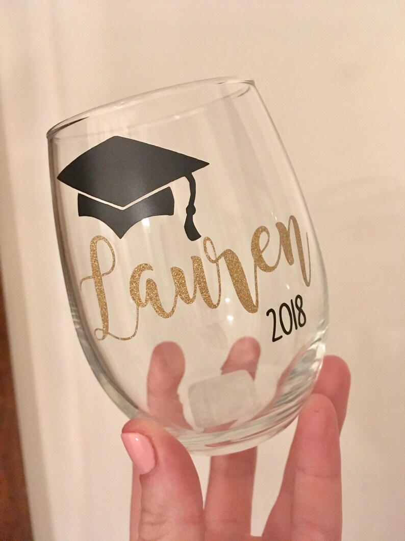 May include: A clear glass wine glass with a black graduation cap and the name "Lauren" in gold glitter lettering and the year "2018" in black lettering.