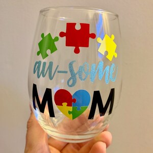 May include: A clear stemless wine glass with the words "au-some MOM" in black and light blue glitter lettering. The design includes colorful puzzle pieces in green, red, yellow, and blue, forming a heart shape.