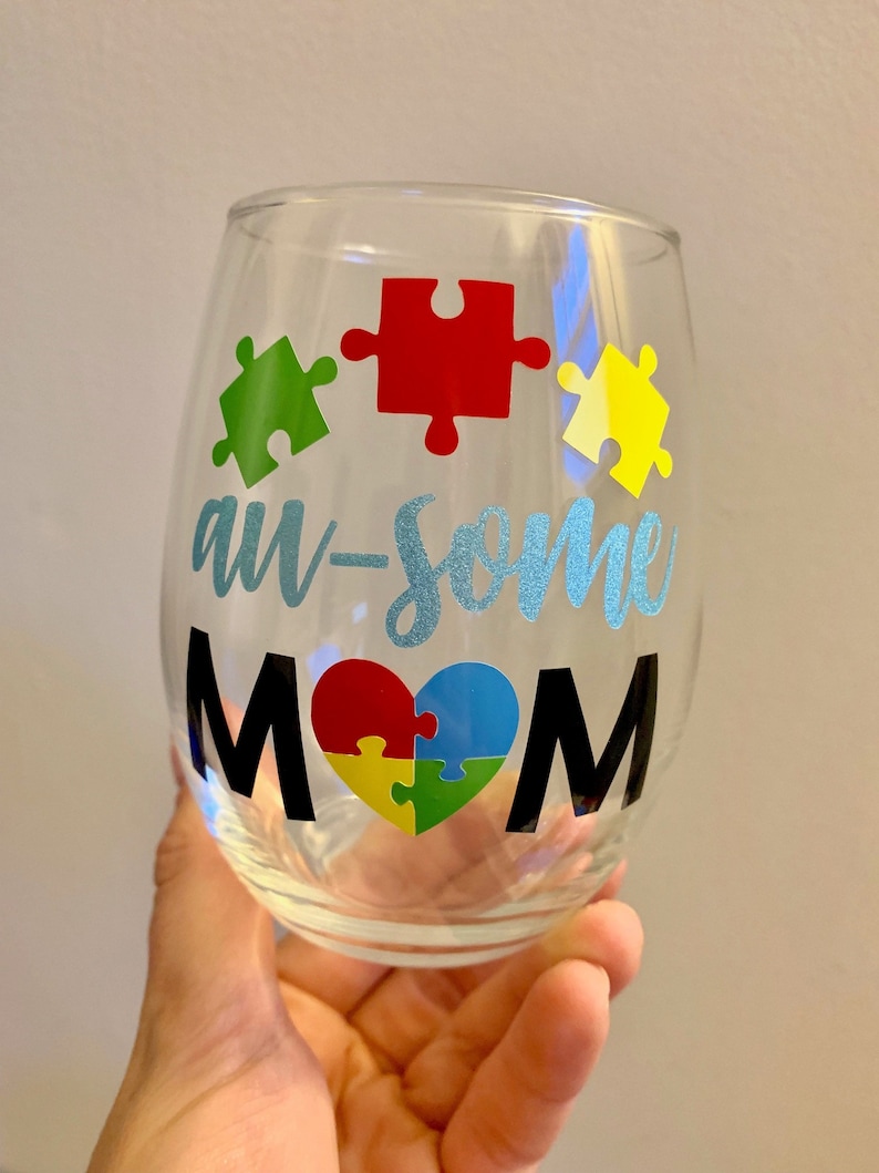 May include: A clear stemless wine glass with the words "au-some MOM" in black and light blue glitter lettering. The design includes colorful puzzle pieces in green, red, yellow, and blue, forming a heart shape.