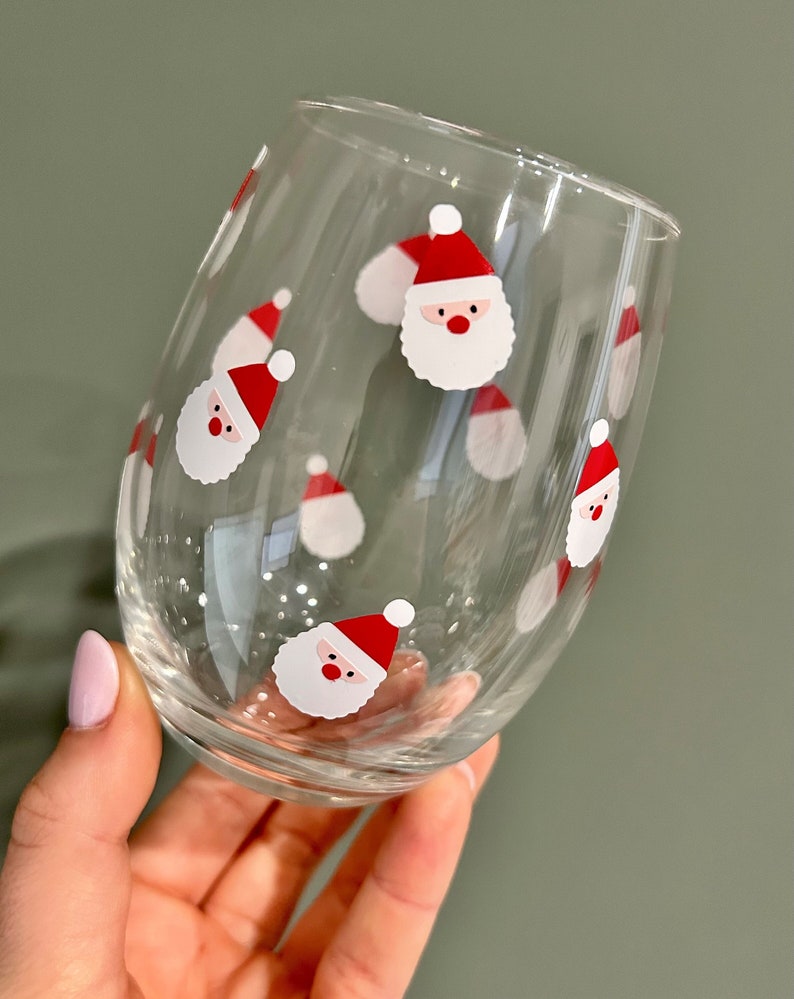 May include: A clear glass wine glass with a festive design featuring multiple red and white Santa Claus faces.