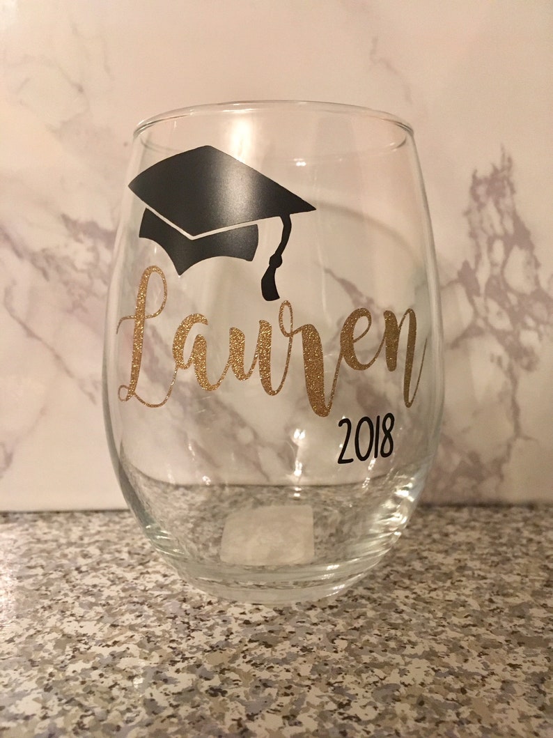 May include: A clear glass wine glass with a black graduation cap and the name "Lauren" in gold glitter and the year "2018" in black.