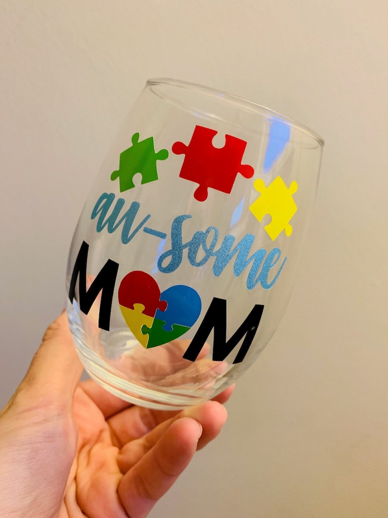 May include: A clear stemless wine glass with the words "aw-some MOM" in black and blue lettering. The design includes colorful puzzle pieces and a heart-shaped puzzle. The glass is held in a hand.