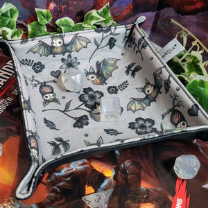 Könnte beinhalten: Ein grauer Stoff-Würfeltablett mit schwarzem Rand. Das Tablett hat ein sich wiederholendes Muster aus Fledermäusen, Blumen und Ranken. Das Tablett ist mit klaren Würfeln gefüllt.