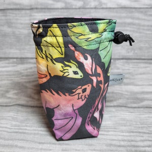 Könnte beinhalten: Eine schwarze Kordeltasche mit einem bunten Drachenmuster. Die Tasche ist aus Stoff und hat einen Kordelverschluss. Die Drachen sind in den Farben Rosa, Gelb, Grün und Blau gehalten.