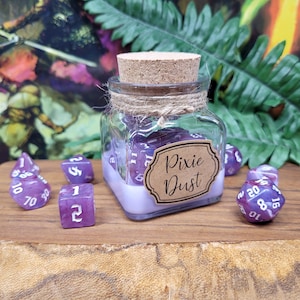 Op de afbeelding: Een glazen pot gevuld met paarse dobbelstenen, met het label "Pixie Dust", staat op een houten oppervlak. De pot is afgesloten met een kurk en touw. Verschillende dobbelstenen liggen verspreid rond de pot, met verschillende vormen en getallen.