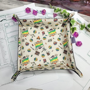 Könnte beinhalten: Ein quadratisches, cremefarbenes Würfeltablett mit schwarzem Rand, das ein sich wiederholendes Muster aus Würfeln, Schwertern, Schilden und Bannern mit der Aufschrift "Roll with Pride" in Regenbogenfarben zeigt. Lila Würfel und D&D-Charakterbögen liegen daneben.