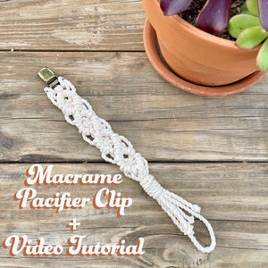 May include: A white macrame pacifier clip with a gold metal clip. The clip is on a wooden surface. The text "Macrame Pacifier Clip + Video Tutorial" is written in orange letters on the surface.
