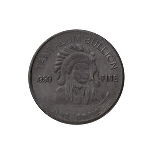 Op de afbeelding: Een zilveren munt met de woorden "Tantalum Bullion .999 Fine One Gram" rond de rand. Het midden van de munt toont een Indiaans hoofd met een hoofdtooi.