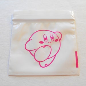 Op de afbeelding: Een witte plastic zak met een roze cartoonfiguur erop gedrukt. Het figuur heeft grote ogen en een kleine mond. De zak is afgesloten met een ritssluiting.