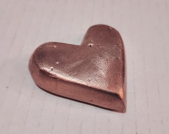 Puede incluir: Un objeto en forma de corazón de color cobre con una superficie texturizada. El corazón es sólido y parece ser de metal. El objeto está sobre una superficie blanca.