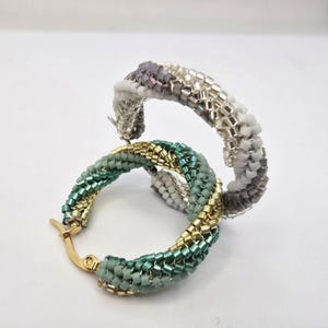 May include: Two hoop earrings crafted from small, woven seed beads. One earring features a color palette of white, grey, and silver beads. The second earring displays a combination of metallic gold, sage green, and teal beads with a gold-tone metal clasp.