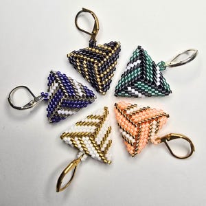 Könnte beinhalten: Fünf dreieckige Ohrringe aus kleinen Glasperlen in verschiedenen Farbkombinationen, darunter Schwarz und Gold, Grün und Weiß, Violett und Silber, Weiß und Gold sowie Pfirsich und Gold. Jeder Ohrring verfügt über einen metallenen Klappbügel.