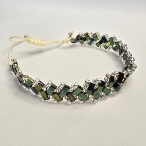 Può includere: Un bracciale con un motivo intrecciato di perline rettangolari iridescenti verde scuro e viola, arricchito da piccole perline di vetro trasparente. Il gioiello presenta una chiusura a cordino regolabile chiaro con nodi scorrevoli.