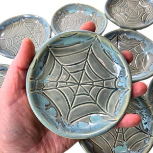 May include: A collection of small, round ceramic dishes with a spiderweb design. The dishes are primarily gray with a turquoise blue rim. The spiderweb pattern is raised, creating a textured surface.