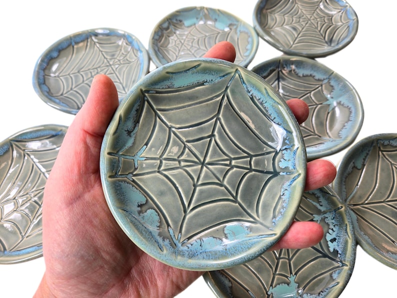 May include: A collection of small, round ceramic dishes with a spiderweb design. The dishes are primarily gray with a turquoise blue rim. The spiderweb pattern is raised, creating a textured surface.