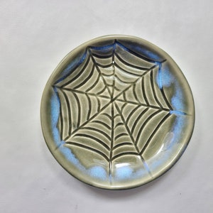 May include: A round, ceramic plate with a spiderweb design. The plate has a gray base color with a blue iridescent glaze around the rim. The spiderweb is a darker gray color.