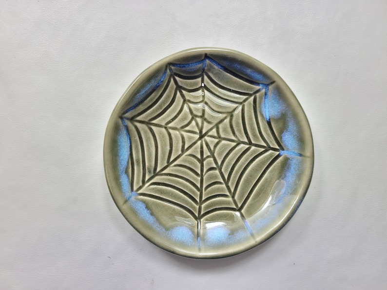 May include: A round, ceramic plate with a spiderweb design. The plate has a gray base color with a blue iridescent glaze around the rim. The spiderweb is a darker gray color.