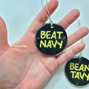 May include: Two black round ceramic ornaments with yellow lettering. One ornament says "BEAT NAVY" and the other says "BEAN TAVY".