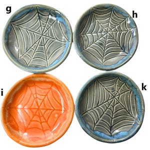 May include: Four ceramic bowls with a spiderweb design. Three bowls are gray with a blue rim, and one is orange. The spiderweb design is raised, creating a textured surface. These bowls are perfect for Halloween or spooky-themed decor.