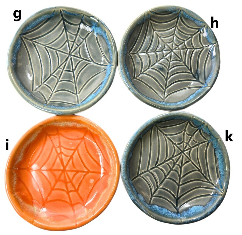 May include: Four ceramic bowls with a spiderweb design. Three bowls are gray with a blue rim, and one is orange. The spiderweb design is raised, creating a textured surface. These bowls are perfect for Halloween or spooky-themed decor.
