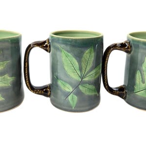 May include: Three ceramic mugs with brown handles and green leaf designs. The mugs are a dark green color with light green leaves.