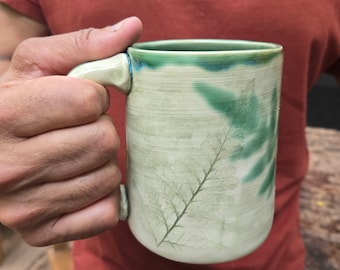 May include: A ceramic mug with a green glaze and a leaf design. The mug has a handle and is made of a light-colored clay.
