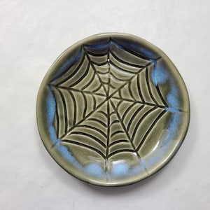 May include: A ceramic plate with a spiderweb design. The plate is round with a gray base and a blue rim. The spiderweb is black and covers the center of the plate. The plate is likely handmade and suitable for Halloween.