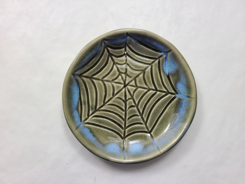 May include: A ceramic plate with a spiderweb design. The plate is round with a gray base and a blue rim. The spiderweb is black and covers the center of the plate. The plate is likely handmade and suitable for Halloween.