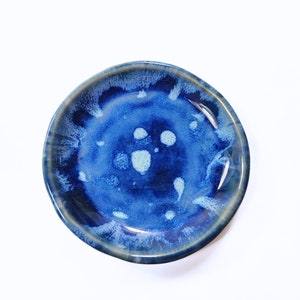 May include: A small, round, blue ceramic dish with white speckles. The dish is glazed and has a slightly uneven surface.