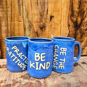 May include: Three blue ceramic mugs with white lettering. The mugs say "Practice Gratitude", "Be Kind", and "Be the Change".