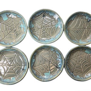 May include: Six ceramic plates with a spiderweb design. The plates are round with a gray web pattern in the center and a turquoise blue rim. The plates are arranged in two rows of three.
