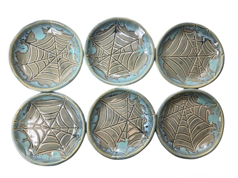 May include: Six ceramic plates with a spiderweb design. The plates are round with a gray web pattern in the center and a turquoise blue rim. The plates are arranged in two rows of three.