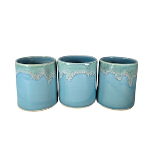 May include: Three cylindrical ceramic cups featuring a smooth, light blue base glaze that transitions into a textured, frothy white and pale blue drip pattern near the top rim. Each cup has a consistent shape and a glossy finish throughout the exterior.