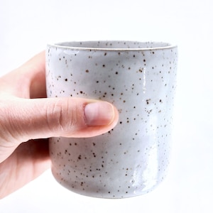 May include: A speckled, off-white ceramic tumbler. The cylindrical cup has a slightly rounded base and a smooth rim. The surface is covered in small, dark brown speckles. The cup is held by a hand against a white background.