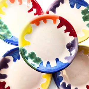 May include: A collection of five ceramic plates with a white center and a colorful, dripped paint design around the edges. The colors include red, orange, yellow, green, blue, and purple. The plates are arranged in a circular pattern.