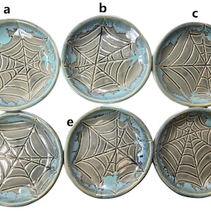 May include: Six ceramic plates with a spiderweb design. The plates are round with a gray center and a turquoise blue rim. The spiderweb design is raised and textured, adding depth to the plates. These plates are ideal for Halloween or spooky-themed decor.