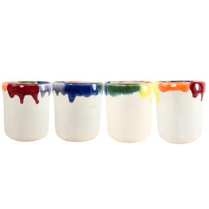 May include: Four white ceramic cups with colorful dripped paint around the rims. The colors include red, blue, green, yellow, and orange. The cups are cylindrical and have a smooth, glossy finish. They are arranged in a row against a white background.
