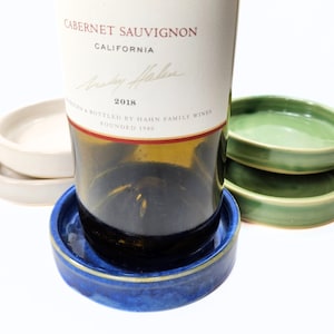May include: A bottle of Cabernet Sauvignon wine rests in a blue ceramic coaster with a gold rim. Several other ceramic coasters in cream and green are stacked nearby. The wine bottle label reads "CABERNET SAUVIGNON CALIFORNIA 2018".